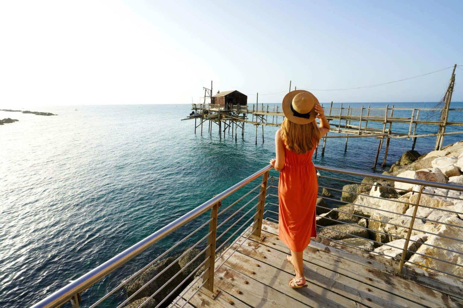 Romantic sunset in Italy. Full length back view of young woman with hat and dress enjoying view of Trabucco an old fishing machine on Adriatic Sea, Italy.